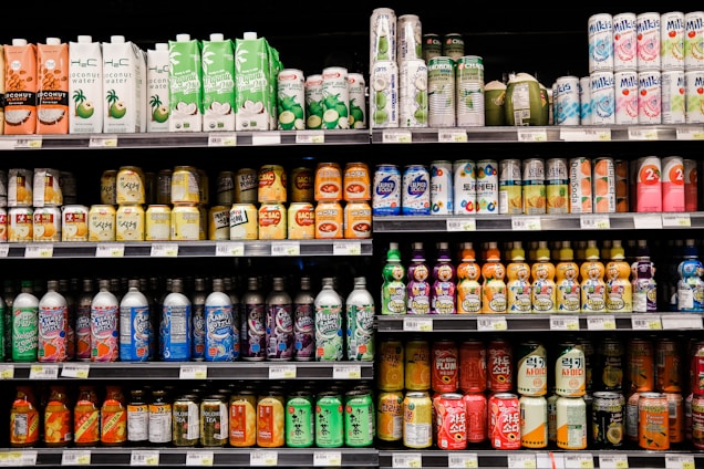A grocery store shelf filled with a wide variety of colorful beverages in cans and bottles. The products include coconut water, carbonated soft drinks, teas, and fruit-flavored drinks. The packaging features vibrant colors and different languages.