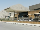 A modern building with an angular roof and surrounding fence is positioned in front of a rocky mountain landscape. Multiple vehicles are parked nearby, with a visible flag featuring red, green, white, and black stripes.