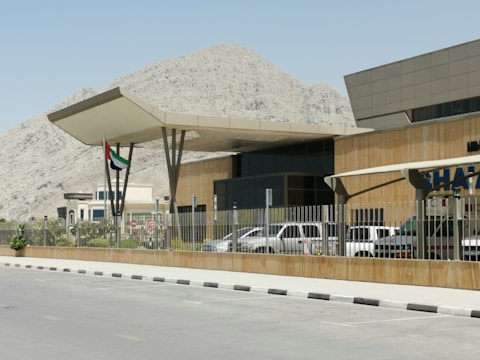 A modern building with an angular roof and surrounding fence is positioned in front of a rocky mountain landscape. Multiple vehicles are parked nearby, with a visible flag featuring red, green, white, and black stripes.