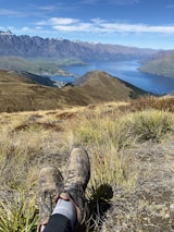 A pair of worn brown hiking shoes rest on a grassy hill overlooking a scenic view of a large, calm lake surrounded by mountainous terrain. Snow-capped peaks are visible in the distance under a clear blue sky, while a town can be seen at the water's edge.