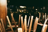 A dimly lit interior of a restaurant or bar with wooden chairs stacked upside down on tables, suggesting closure or preparation for cleaning. The background contains blurred images of shelves and bar paraphernalia.