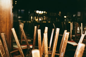A dimly lit interior of a restaurant or bar with wooden chairs stacked upside down on tables, suggesting closure or preparation for cleaning. The background contains blurred images of shelves and bar paraphernalia.