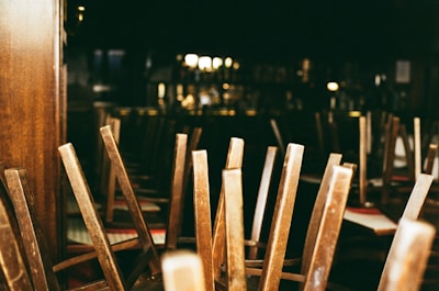 A dimly lit interior of a restaurant or bar with wooden chairs stacked upside down on tables, suggesting closure or preparation for cleaning. The background contains blurred images of shelves and bar paraphernalia.