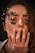 A close-up portrait of a person with intricate makeup art on their face, featuring black and gold embellishments, pearls, and small star-shaped decals. The person's long nails are painted with a French manicure style, and they have a nose ring. The lighting highlights the shimmering decorations around the eyes.