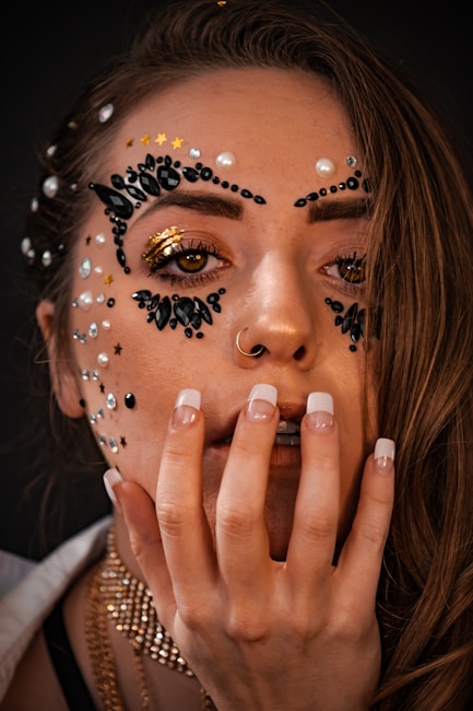 A close-up portrait of a person with intricate makeup art on their face, featuring black and gold embellishments, pearls, and small star-shaped decals. The person's long nails are painted with a French manicure style, and they have a nose ring. The lighting highlights the shimmering decorations around the eyes.
