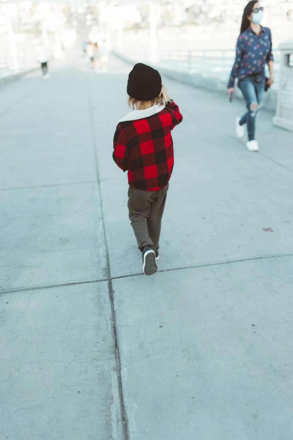 child in red and black plaid shirt and brown pants walking on gray concrete floor