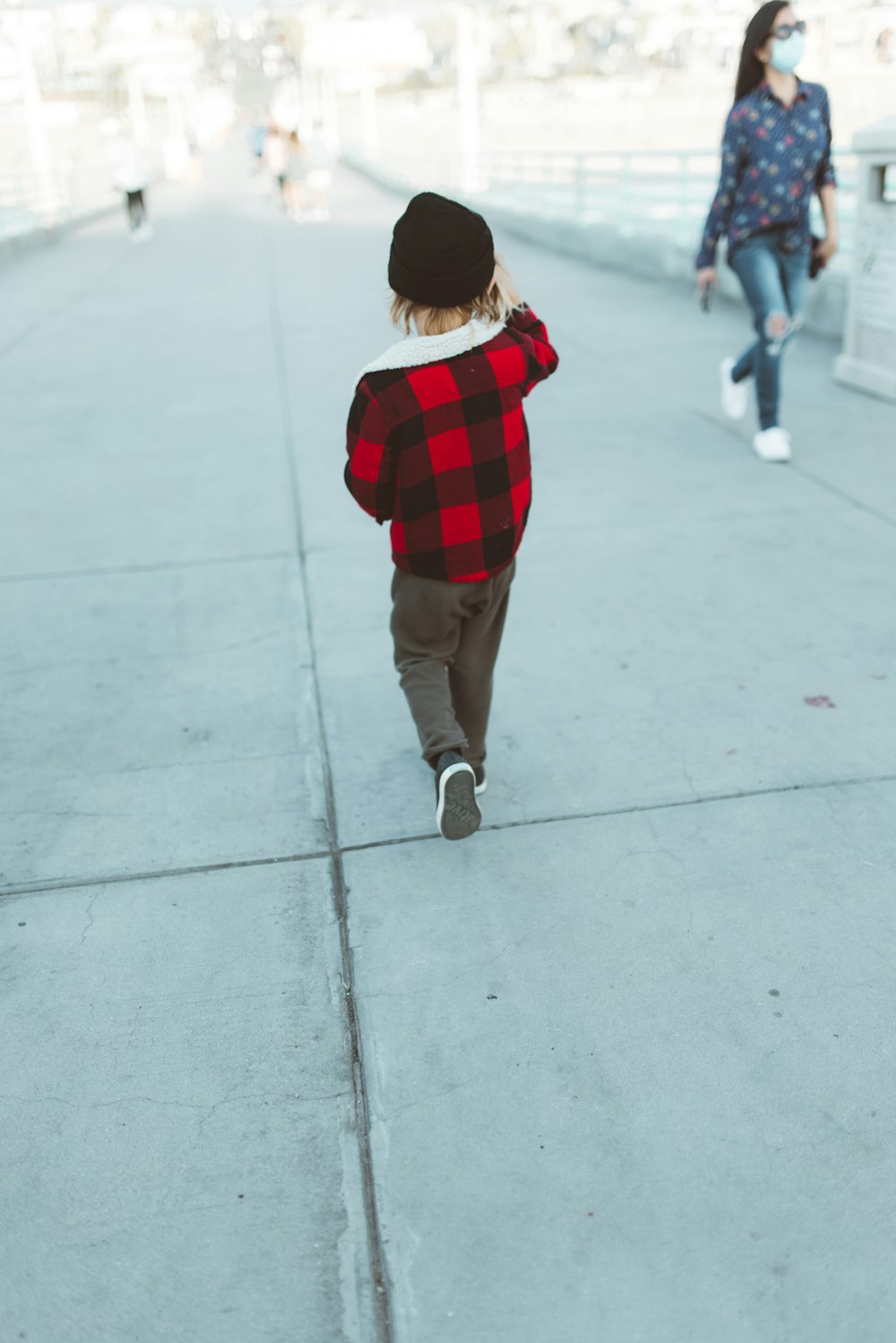 Enfant en chemise à carreaux rouge et noir et pantalon marron marchant sur un sol en béton gris