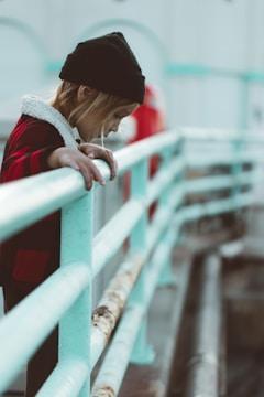 A child wearing a black beanie and a red checkered coat leans over a faded turquoise railing, appearing deep in thought. The background is softly blurred, emphasizing the child's attentive expression.