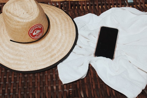 A straw hat with a red circular logo reading 'Quiksilver Bali Surf Academy' is placed on a wicker surface next to a white towel. Beside the towel, there is a smartphone with a black screen.