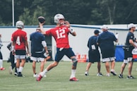 A Patriots player throwing a precise pass, framed by the gray and red team colors.