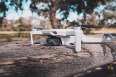 A small drone rests on a wooden surface, surrounded by a natural, blurred background of trees and grass. The drone has a sleek, compact design with visible propellers and a camera attached to its front.