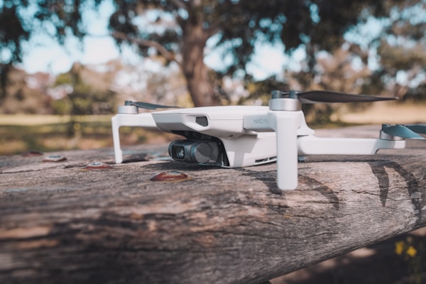 A small drone rests on a wooden surface, surrounded by a natural, blurred background of trees and grass. The drone has a sleek, compact design with visible propellers and a camera attached to its front.