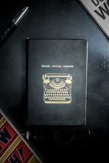 A notebook with a black cover featuring a gold illustration of a typewriter and the words 'think, write, create' on the front. It is placed on a dark surface accompanied by a black pen and a partial view of a newspaper or magazine.