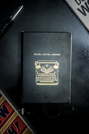 A notebook with a black cover featuring a gold illustration of a typewriter and the words 'think, write, create' on the front. It is placed on a dark surface accompanied by a black pen and a partial view of a newspaper or magazine.