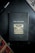 A notebook with a black cover featuring a gold illustration of a typewriter and the words 'think, write, create' on the front. It is placed on a dark surface accompanied by a black pen and a partial view of a newspaper or magazine.