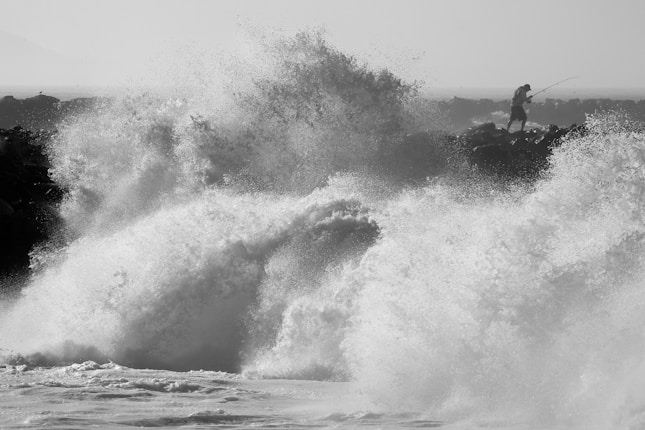A person is standing on rocky outcrops at the ocean's edge, holding a fishing rod amidst large crashing waves. The water is forcefully splashing, creating a dramatic scene filled with motion.