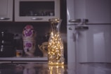 A kitchen setting features a glass carafe filled with warm, glowing fairy lights on a reflective surface. In the blurred background, there is a refrigerator and objects including a ceramic jar with floral patterns.