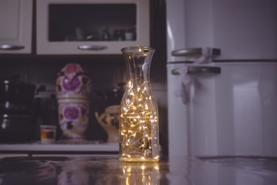 A kitchen setting features a glass carafe filled with warm, glowing fairy lights on a reflective surface. In the blurred background, there is a refrigerator and objects including a ceramic jar with floral patterns.