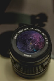 A camera lens with a cosmic scene reflecting or appearing on the glass surface. The scene shows a colorful nebula with stars, blending elements of astrophotography and optical equipment.