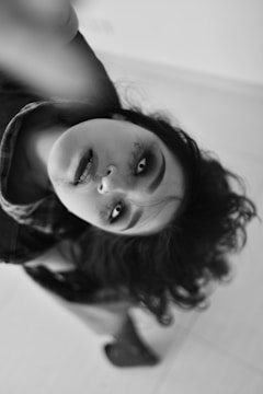 A black and white photograph captures a person with dramatic makeup and intense eye expression. Their head is tilted, and hair cascades around their face, creating a dynamic and slightly unsettling visual effect. The background is minimal, emphasizing the subject's features and expression.
