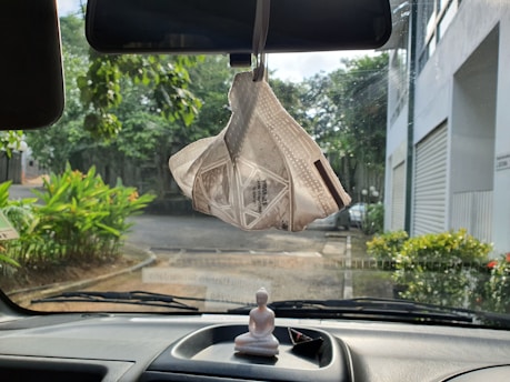 A white face mask hangs from the rearview mirror inside a vehicle with a dashboard that includes a small Buddha statue and a triangular paper object. Outside the windshield, there is a view of greenery, a road, and buildings.