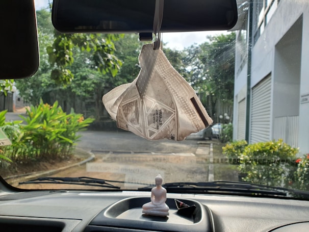 A white face mask hangs from the rearview mirror inside a vehicle with a dashboard that includes a small Buddha statue and a triangular paper object. Outside the windshield, there is a view of greenery, a road, and buildings.