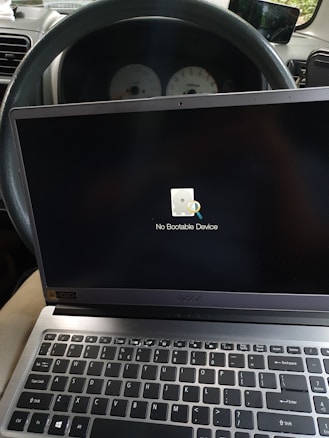 An open laptop is displaying a message 'No Bootable Device' on its screen. The laptop is placed on the driver’s seat inside a car, with the steering wheel and car dashboard visible in the background.