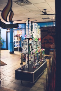 A display of various glass bongs is arranged neatly on a table in a shop with blue walls and tiled flooring. The background includes shelves showcasing different products, under soft indoor lighting. The setting appears organized and colorful.