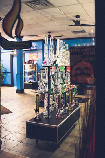 A display of various glass bongs is arranged neatly on a table in a shop with blue walls and tiled flooring. The background includes shelves showcasing different products, under soft indoor lighting. The setting appears organized and colorful.