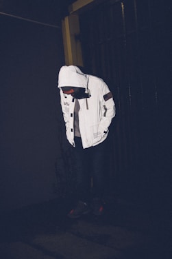 A person is wearing a white hooded jacket with various patches and logos. The person is looking down in a dark environment, with shadows around and minimal lighting highlighting the jacket.