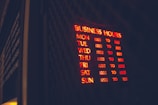 A glowing neon sign displaying business hours mounted on a dark background, with days of the week listed alongside corresponding opening and closing times in red and yellow light. The words are clear in the dark environment, creating a vibrant and inviting appearance.