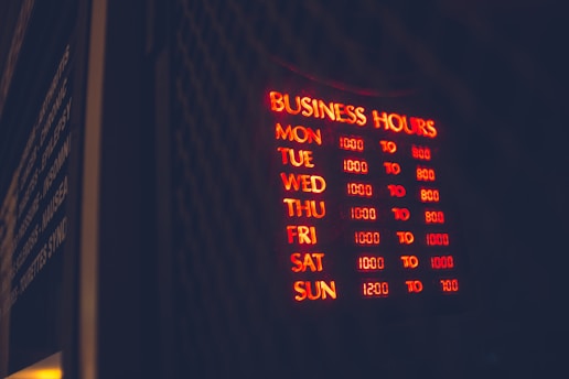 A glowing neon sign displaying business hours mounted on a dark background, with days of the week listed alongside corresponding opening and closing times in red and yellow light. The words are clear in the dark environment, creating a vibrant and inviting appearance.
