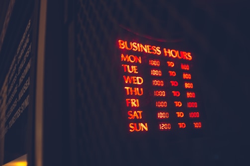A glowing neon sign displaying business hours mounted on a dark background, with days of the week listed alongside corresponding opening and closing times in red and yellow light. The words are clear in the dark environment, creating a vibrant and inviting appearance.