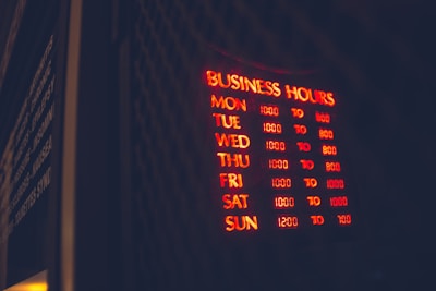 A glowing neon sign displaying business hours mounted on a dark background, with days of the week listed alongside corresponding opening and closing times in red and yellow light. The words are clear in the dark environment, creating a vibrant and inviting appearance.