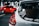 A white car is seen in the foreground with various stickers on the rear window, including a warning not to touch the car and a graphic of a stick figure. In the background, a bright red car is parked in a dimly lit garage.