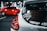 A white car is seen in the foreground with various stickers on the rear window, including a warning not to touch the car and a graphic of a stick figure. In the background, a bright red car is parked in a dimly lit garage.