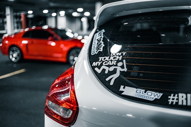 A white car is seen in the foreground with various stickers on the rear window, including a warning not to touch the car and a graphic of a stick figure. In the background, a bright red car is parked in a dimly lit garage.
