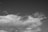 A black and white photograph featuring a large cloud formation dominating the sky. In the center, there is a small silhouette of an airplane flying amidst the clouds.