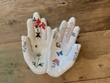 A ceramic dish shaped like a pair of hands, with intricate floral and butterfly designs painted on it. The fingers and palms are adorned with various colorful flowers and a butterfly, all outlined in gold lines. The dish is placed on a rustic wooden surface.