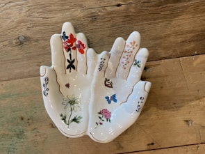 A ceramic dish shaped like a pair of hands, with intricate floral and butterfly designs painted on it. The fingers and palms are adorned with various colorful flowers and a butterfly, all outlined in gold lines. The dish is placed on a rustic wooden surface.