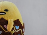 A plush toy with a yellow body and a face featuring minimalistic eyes and a mouth. It has a brown accessory resembling a wrapper with colorful heart patterns and wavy lines around it.