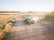 A sunny rural farming scene features a green tractor transferring crops into a large trailer. Dust is being kicked up from the harvesting process in the middle of a vast open field surrounded by golden crops. A small truck is parked near hay bales, and trees line the horizon.