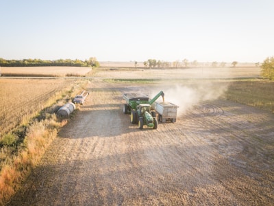 A sunny rural farming scene features a green tractor transferring crops into a large trailer. Dust is being kicked up from the harvesting process in the middle of a vast open field surrounded by golden crops. A small truck is parked near hay bales, and trees line the horizon.