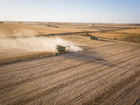 Two green combine harvesters are working in a vast, open field, leaving trails of dust behind them as they harvest crops. The landscape is flat and expansive, characterized by golden-brown tones of harvested farmland extending to the horizon under a clear blue sky.