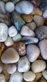A collection of smooth, polished stones in various shapes and sizes, featuring shades of white, brown, gray, and green. Each stone has a unique texture and pattern, contributing to an overall natural and earthy appearance.