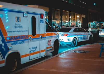 An ambulance and police cars are parked on the side of a street at night, with vibrant lighting coming from nearby buildings and streetlights. The vehicles are marked with emergency service labels and are seen under neon and street lighting, casting shadows on the pavement.