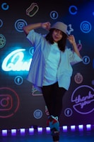 A person wearing a bucket hat and a casual outfit poses playfully. The background features vibrant neon signs and logos on a dark wall, including social media icons.
