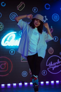 A person wearing a bucket hat and a casual outfit poses playfully. The background features vibrant neon signs and logos on a dark wall, including social media icons.