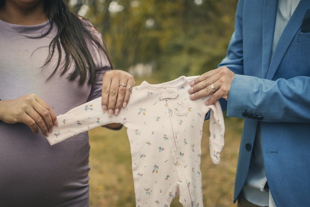 A couple holds up a small baby onesie between them outdoors. The woman on the left is wearing a light purple top, and the man on the right is wearing a blue suit jacket. The background features blurred greenery, indicating a natural setting.
