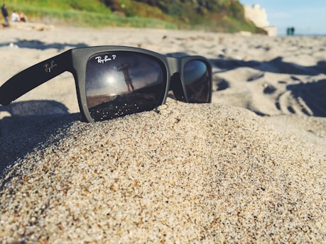 A pair of Ray-Ban sunglasses is placed on a mound of sand at a beach. The lenses reflect a person standing with arms outstretched, with the ocean and some greenery visible in the background. The scene suggests a sunny day with clear skies.
